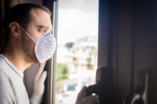 Man with face mask and gloves looking out the window. Stay at home concept.