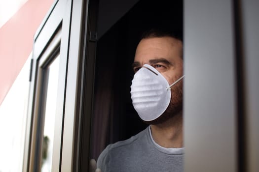 Man with face mask and gloves looking out the window. Stay at home concept.