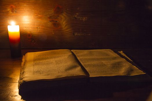 Reading scene in ancient times: an old book leaning on ruined wooden table lighted by a candle on a wooden background