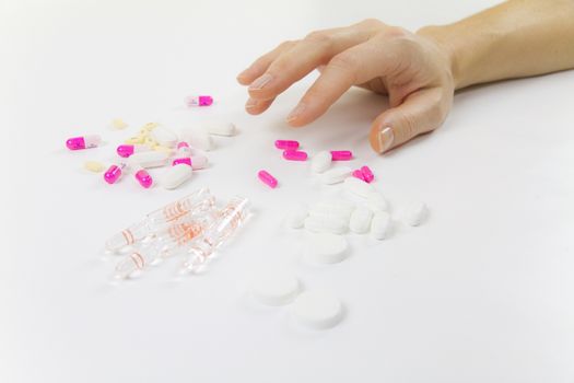 Addiction to medicines and drugs in modern times: close-up shot of a young woman's hand about to grab various tablets and vials on table and white background