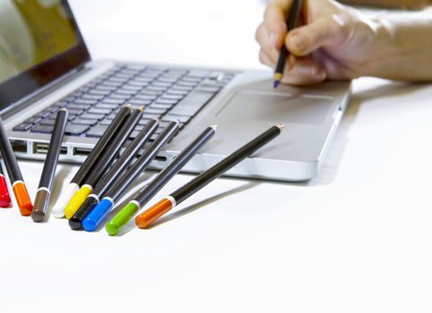 Digital or traditional graphic arts? A woman's hand seems to draw on the touch pad of a laptop computer with lots of colored pencils resting on the side