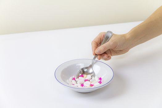 Addiction to medicines and drugs in modern times: a hand of a young woman with a spoonful of tablets taken from a plate full of medicines on white table and background