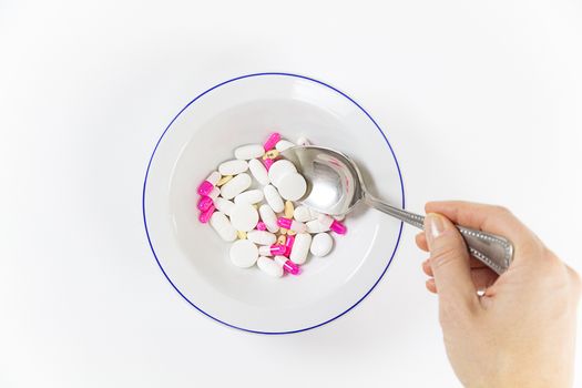 Addiction to medicines and drugs in modern times: a hand of a young woman with a spoonful of tablets taken from a plate full of medicines on white table and background