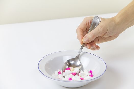 Addiction to medicines and drugs in modern times: a hand of a young woman with a spoonful of tablets taken from a plate full of medicines on white table and background