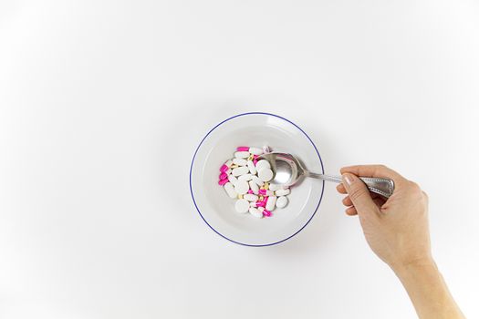 Addiction to medicines and drugs in modern times: a hand of a young woman with a spoonful of tablets taken from a plate full of medicines on white table and background