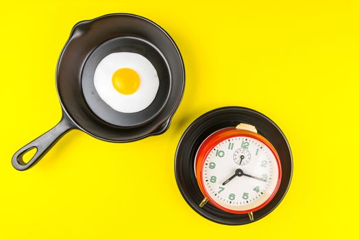 Frying pan with fried egg and alarm clock in a black plate isolated on a yellow background viewed from above.Concept of breakfast at the race against time.