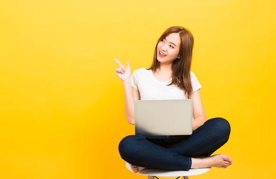 Asian happy portrait beautiful cute young woman teen smiling sitting crossed legs on chair with laptop computer point finger side looking to side isolated, studio shot yellow background with copy space