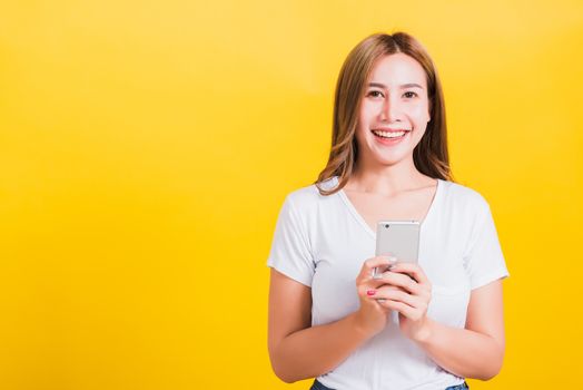 Asian Thai portrait happy beautiful cute young woman smiling stand wear t-shirt lift hand celebrating winning with smart mobile phone looking to camera isolated, on yellow background with copy space