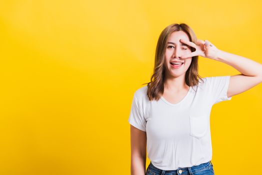 Asian Thai happy portrait beautiful cute young woman smile standing wear t-shirt showing finger making v-sign symbol near eye looking to camera, isolated studio shot yellow background with copy space