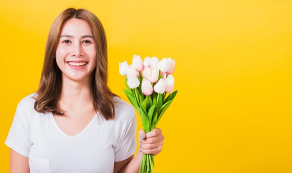 Portrait Asian Thai beautiful happy young woman smiling, screaming excited hold flowers tulips bouquet in hands and looking to camera, studio shot isolated on yellow background, with copy space