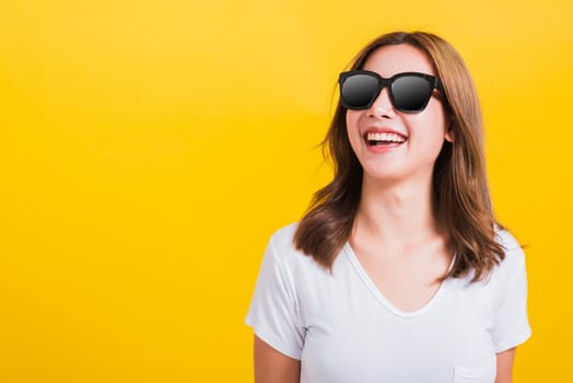 Portrait Asian Thai beautiful young woman happy smiling white teeth with sunglasses and looking side space, studio shot isolated on yellow background, with copy space