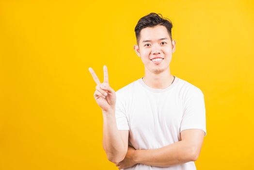 Portrait happy Asian handsome young man smiling standing wearing white t-shirt he showing fingers doing victory V sign or number two, looking to camera, studio shot isolated yellow background