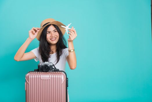 Traveler tourist happy Asian beautiful young woman, holidays travel concept, her holding fly toy aircraft her have suitcase bag and photo mirrorless camera, shoot photo in studio on blue background