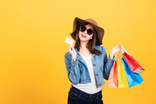 Asian happy portrait beautiful young woman teen smiling standing with sunglasses excited using credit card enjoys shopping and hold shopping bags multi color isolated, studio short yellow background