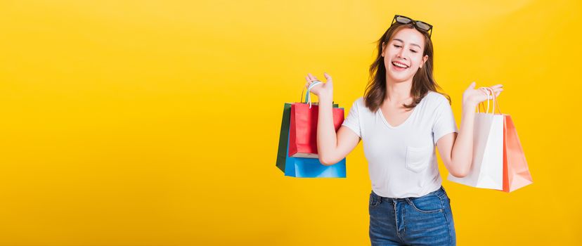 Asian Thai portrait happy beautiful cute young woman smiling stand with sunglasses excited holding shopping bags multi color looking down, studio shot isolated yellow background with copy space