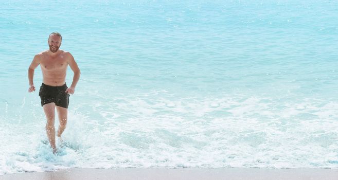 Happy smiling man with perfect body running from sea waves splashing water. Vacations on sea shore concept