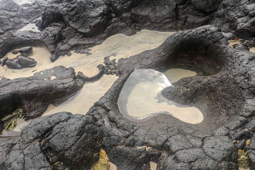 Depressions in a rock with white sand at the bottom, flooded with sea water. Small ponds excavated in lava bed on Indian Ocean coast, Bali, Indonesia. The tide dug out small lakes in the black rock.