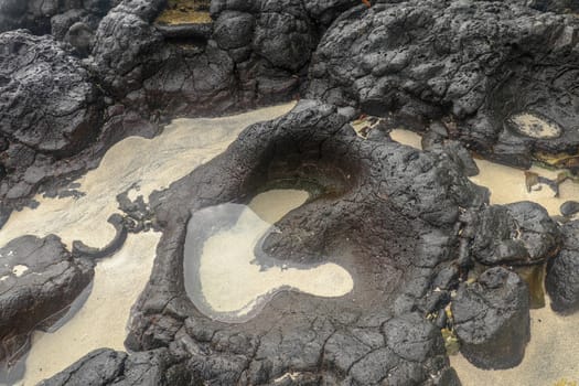 Depressions in a rock with white sand at the bottom, flooded with sea water. Small ponds excavated in lava bed on Indian Ocean coast, Bali, Indonesia. The tide dug out small lakes in the black rock.