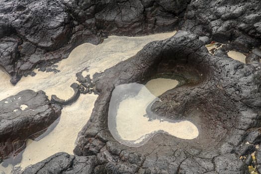 Depressions in a rock with white sand at the bottom, flooded with sea water. Small ponds excavated in lava bed on Indian Ocean coast, Bali, Indonesia. The tide dug out small lakes in the black rock.