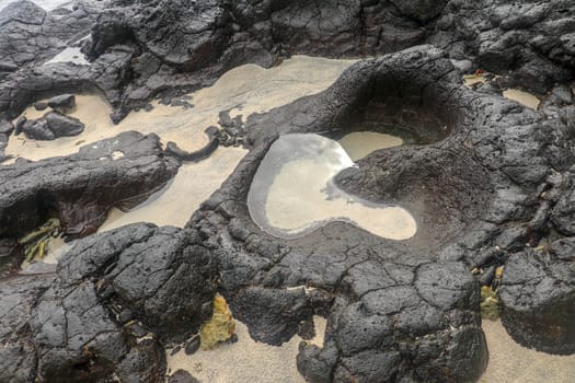 Depressions in a rock with white sand at the bottom, flooded with sea water. Small ponds excavated in lava bed on Indian Ocean coast, Bali, Indonesia. The tide dug out small lakes in the black rock.