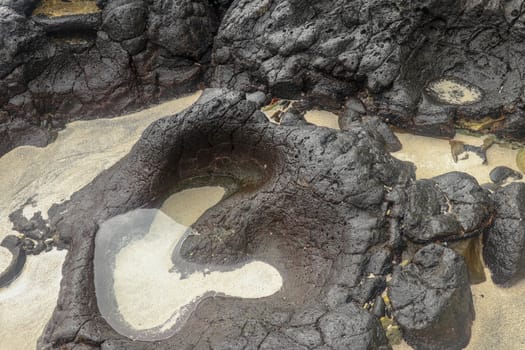 Depressions in a rock with white sand at the bottom, flooded with sea water. Small ponds excavated in lava bed on Indian Ocean coast, Bali, Indonesia. The tide dug out small lakes in the black rock.