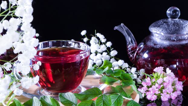 A mug of red tea and a teapot in white hibiscus flowers and green leaves of medicinal tea on a wooden stand.Zen tea ceremony. Photo of red herbal Indian healing tea.Elegant mugs with a relaxing drink