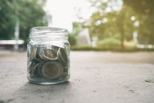 Coins in Glass Jar in Park with Light Leak. Translation Text on Coin is "Thailand, 10 Bath". (Baht is Currency of Thailand)