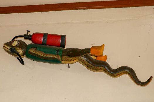 Statue of gecko with diving equipment on the wall of diving center. 3D gecko as a diver. Lizard figurine with green jacket, red diving tank and yellow fins. A funny accessory in the diving base area.