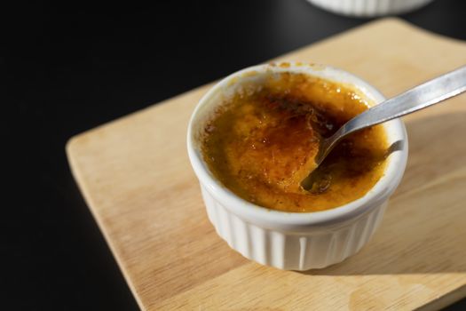 Homemade creme brulee in white ramekin  with sugar burn top up cracking with spoon on black background with copy space. French dessert creme brulee