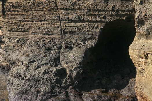 Dark rock of volcanic lava on the Indian Ocean coast of Bali. A small cave excavated into a cliff wall by a sea surf. Sun rays illuminate the rough wall of a rock cliff.