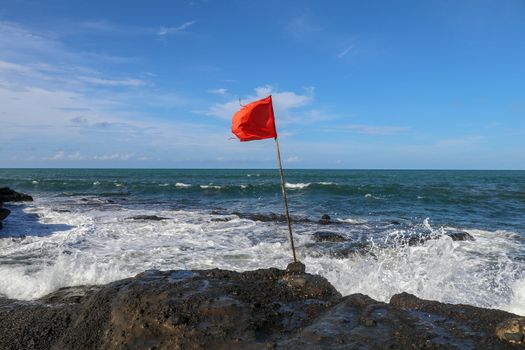 Red flag on a wooden flagpole flies on coast of Indian Ocean. Sign warning of danger of big breakers. Rock cliff at Sacred Balinese temple Tanah Lot. Waves are breaking on rock cliffs of black lava.