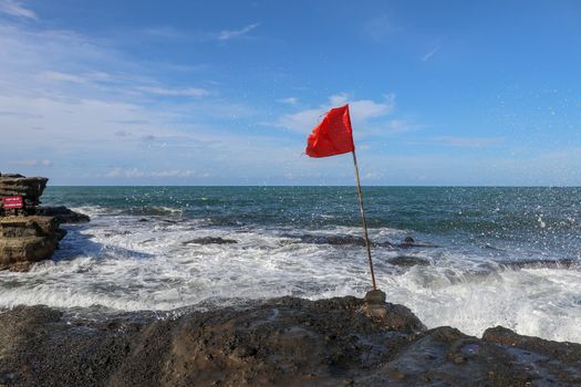 Red flag on a wooden flagpole flies on coast of Indian Ocean. Sign warning of danger of big breakers. Rock cliff at Sacred Balinese temple Tanah Lot. Waves are breaking on rock cliffs of black lava.