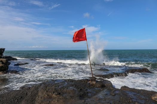 Red flag on a wooden flagpole flies on coast of Indian Ocean. Sign warning of danger of big breakers. Rock cliff at Sacred Balinese temple Tanah Lot. Waves are breaking on rock cliffs of black lava.