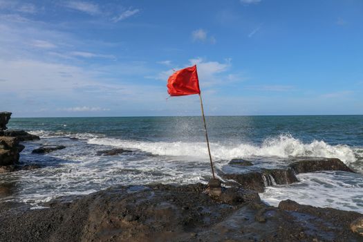 Red flag on a wooden flagpole flies on coast of Indian Ocean. Sign warning of danger of big breakers. Rock cliff at Sacred Balinese temple Tanah Lot. Waves are breaking on rock cliffs of black lava.