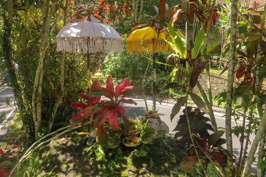A sacred place at the driveway to Hindu temple Batukaru on Bali island in Indonesia. Sacrifices to God from believers. Closeup of stones, yellow and white decorative umbrelas among tropical plants.