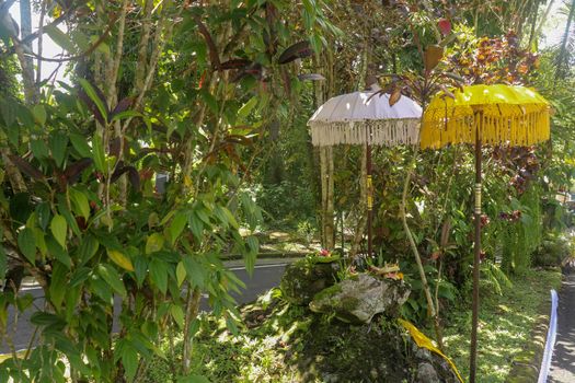 A sacred place at the driveway to Hindu temple Batukaru on Bali island in Indonesia. Sacrifices to God from believers. Closeup of stones, yellow and white decorative umbrelas among tropical plants.