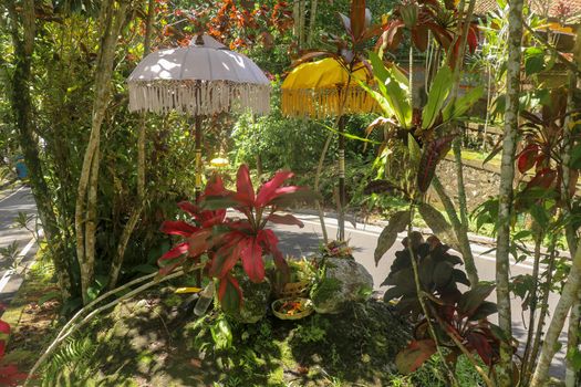 A sacred place at the driveway to Hindu temple Batukaru on Bali island in Indonesia. Sacrifices to God from believers. Closeup of stones, yellow and white decorative umbrelas among tropical plants.