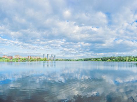 Large blue lake and sky with beautiful clouds with green forest, houses and skyscrapers on the shore. Panorama landscape