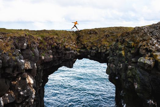 Tourist jumps over a natural rock bridge connecting basalt cliffs in Arnarstapi, Iceland, with Atlantic Ocean in the background.