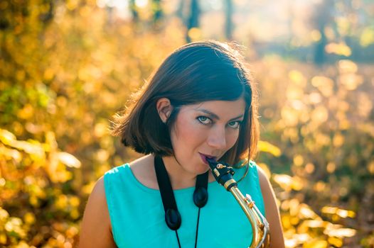 cheerful young woman with dark hair in blue clothes plays the saxophone in autumn yellow park