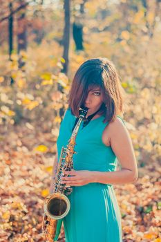 charming young brunette girl in a long blue dress plays the alto saxophone in a yellow autumn park