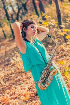 cute young dark-haired girl with saxophone posing in a long blue dress in a yellow autumn forest, raising hands to head