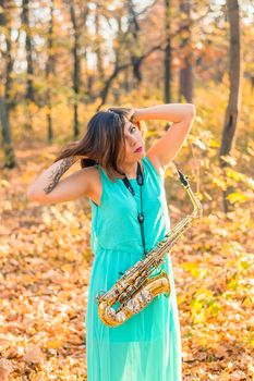 lovely brunette girl in a long blue dress and alto saxophone posing in a yellow autumn park, raising hands to head