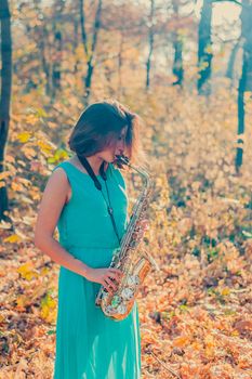 cute young brunette girl in a long blue dress plays the yellow saxophone in the autumn forest