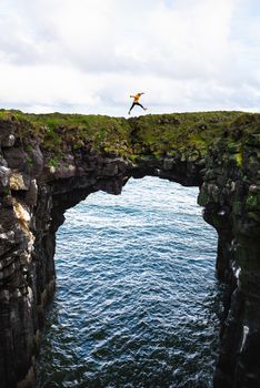 Tourist jumps over a natural rock bridge connecting basalt cliffs in Arnarstapi, Iceland, with Atlantic Ocean in the background.