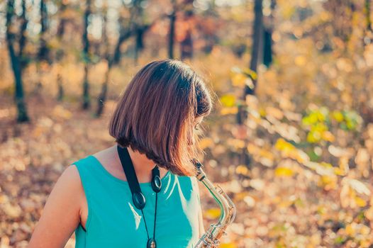 sideways from close up a brunette woman in a blue dress plays the saxophone in a yellow autumn park