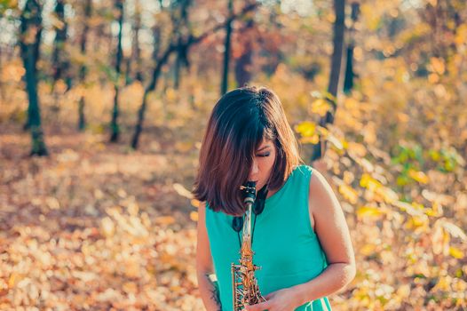 teenager girl with black hair in a blue dress enthusiastically plays the saxophone in the yellow autumn forest