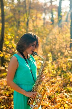 cute black-haired girl in a blue dress plays the yellow saxophone in the autumn forest