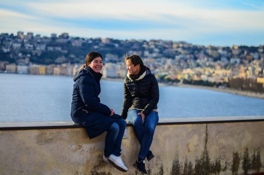 two smiling girls sit near the sea. City ​​in the background. Naples, Italy