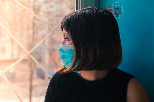 brunette girl in a black dress sits in a surgical  mask sits on a hospital window and looks outside